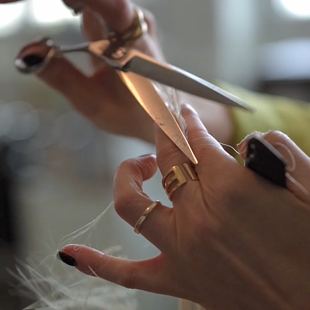 Eine Nahaufnahme zeigt die Hände einer Person, die mit einer Schere Haarsträhnen abschneidet. Die Person trägt mehrere goldene Ringe und schwarzen Nagellack. Die Szene scheint in einem Friseursalon mit sanftem Tageslicht im Hintergrund zu spielen.