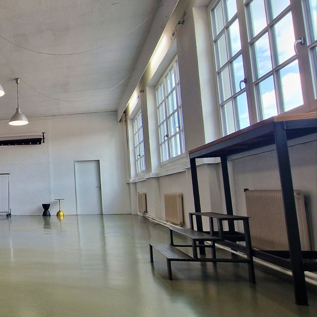 Ein geräumiger, minimalistischer Raum mit hohen Decken, großen Fenstern, die natürliches Licht hereinlassen, einem einfachen Tisch mit Hockern, einem kleinen Beistelltisch; Wände und Boden sind hell.