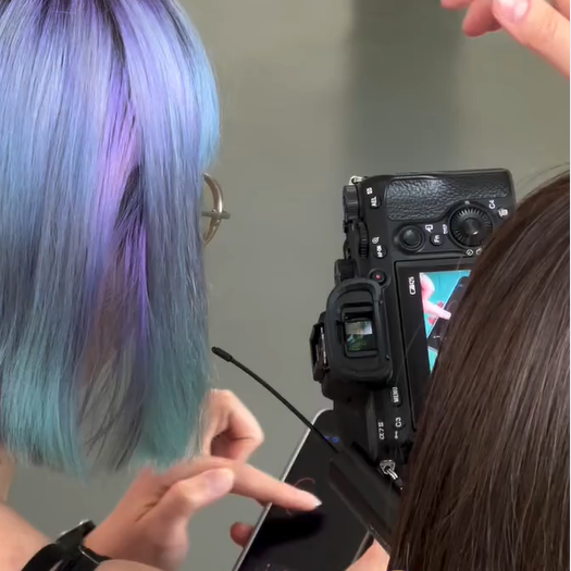 Eine Person mit blau-violetten Haaren zeigt auf einen Smartphone-Bildschirm, während eine andere eine Kamera hält, die eine Fotovorschau zeigt. Beide blicken auf das Kameradisplay.